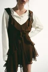 Vestido corto color marrón con escote en pico y tirantes anchos. Presenta un diseño con múltiples volantes en cascada que recorren el frente y el bajo, con un acabado asimétrico.
