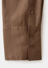 Blusa de ramio color marrón, de diseño recto, con cuello camisero, manga larga con puños abotonados, cierre frontal con botones y detalle de lazo cruzado.