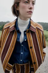 Chaqueta tipo bomber de cuello solapa y manga larga, con cierre frontal de botones y bolsillos de vivo en el delantero. Presenta un diseño a rayas verticales en tonos marrones y beige.