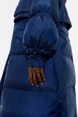 Plumífero largo azul con relleno de pluma y plumón, cuello alto, manga larga, cierre frontal con cremallera, botones de presión y cinturón. Tiene bolsillos laterales y capucha extraíble con pelo a contraste.