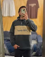 Buzo de polar térmico con diseño de bloques de color en gris y beige. Presenta cuello alto con cierre frontal parcial y bordado en el pecho. Posee puños elásticos y corte holgado.