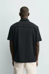 Camisa regular fit negra de manga corta, con cuello bowling y cierre frontal de botonadura. Confeccionada en tejido texturizado con elasticidad y aberturas laterales en el bajo.