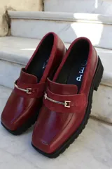 Mocasín de cuero vacuno color bordó con suela track negra de goma. Presenta un diseño de punta cuadrada con un herraje metálico plateado sobre una banda de gamuza en la capellada.