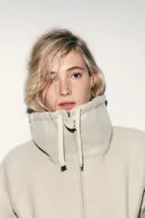 Sudadera tipo bomber de tejido suave, color beige, con cuello alto ajustable mediante cordones, cierre frontal con cremallera, mangas largas con puños elásticos y bolsillos laterales de vivo.