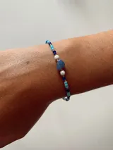 Pulsera elástica compuesta por mostacillas en tonos azules, celestes y blancos, con cuentas blancas facetadas y un dije central de piedra azul en forma de corazón.