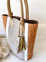 Bolso tipo tote bag de cuero color marrón con textura tipo cocodrilo, detalles en blanco y manijas de cuero color beige. Incluye dos borlas decorativas y correa larga opcional.