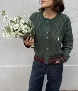 Saco tejido de punto color verde oscuro con diseño de ochos y trenzas. Presenta cierre frontal con botones, cuello redondo y detalles en color bordó en los puños y el ruedo. Incluye un delicado bordado a mano en el pecho.