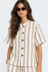 Blusa de manga corta a rayas beige y blancas, con cuello mao y cierre frontal de botones con trabillas (alamares). Cuenta con tajos laterales.