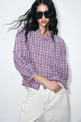 Blusa de manga larga con diseño a cuadros en tonos violeta, rosa y blanco. Presenta cuello redondo, cierre frontal con botones y mangas con frunces en los hombros.