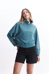 Buzo de felpa color verde azulado (teal) con cuello alto y cierre frontal parcial. Presenta bordados en contraste: un sol pequeño en el pecho y texto en la manga izquierda. Tiene puños y cintura ajustados.