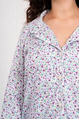Pijama de dos piezas, compuesto por una camisa de manga larga con botones y cuello clásico, estampada con flores pequeñas en tonos lila y verde sobre fondo blanco, y un pantalón liso color lila con cintura elástica.