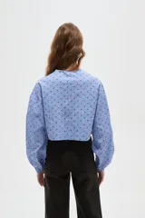 Blusa de corte holgado con estampado de cuadros vichy en azul y blanco, decorada con pequeños corazones rojos bordados. Presenta cuello a la base, mangas largas abullonadas y cierre frontal mediante cordones ajustables.