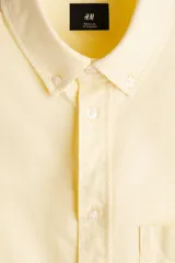 Camisa H&M de algodón Oxford en color amarillo claro, con cuello americano, bolsillo en el pecho y cierre frontal con botones.