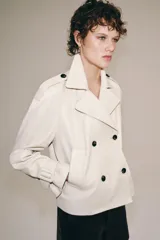 Chaqueta corta de piel 100% color crudo, con cuello de solapa, manga larga con trabillas ajustables y cierre frontal cruzado con botones oscuros.