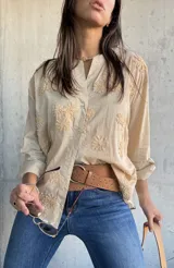 Camisa de color beige con cuello mao y cierre frontal de botones. Presenta un diseño con bordados florales en relieve distribuidos por toda la prenda, mangas largas y corte holgado.