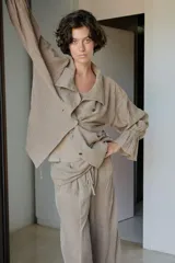 Camisa de lino color tostado con cuello subido, manga larga con puños elásticos y volantes, cintura ajustable con lazadas laterales y cierre frontal cruzado con doble botonadura.