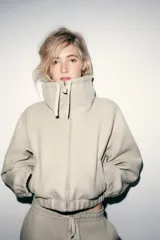 Sudadera tipo bomber de tejido suave, color beige, con cuello alto ajustable mediante cordones, cierre frontal con cremallera, mangas largas con puños elásticos y bolsillos laterales de vivo.