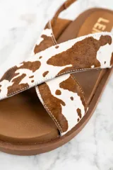 Sandalias con tiras cruzadas de cuero con estampado animal print de vaca en tonos marrón y blanco. Tira trasera con hebilla ajustable. Suela plana color marrón.