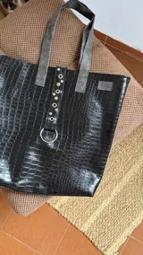 Tote bag de gran tamaño confeccionada en ecocuero negro con textura de reptil. Presenta una tira central decorativa con ojales metálicos y una argolla grande en la parte inferior, además de manijas en color gris.