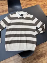 Buzo de tejido de punto a rayas horizontales en tonos beige y marrón, con cuello polo y medio cierre frontal. Presenta puños y ruedo acanalados.
