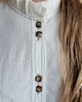 Camisa de manga tres cuartos color beige con cuello con volados y botones frontales. Presenta un detalle de color negro en la parte superior de las mangas.