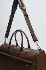 Bolso de viaje color marrón liso con acabado brillante. Cuenta con un compartimento principal con cierre de cremallera, un bolsillo interior con cremallera y un bolsillo frontal con cremallera. Tiene dos asas de mano y una correa de hombro ajustable y extraíble.