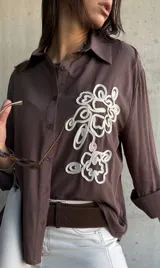 Camisa de manga larga color chocolate, con cuello clásico y cierre frontal de botones. Presenta un bordado decorativo en color crudo en el frente con diseño de formas orgánicas.