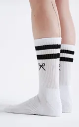 Medias deportivas blancas de caña media, con dos franjas negras horizontales cerca del borde superior y un pequeño logo bordado en negro con forma de lazo o tijeras cruzadas en la parte frontal.
