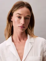 Vestido camisero corto de color blanco, confeccionado en tejido de gasa de algodón. Presenta cuello clásico, cierre frontal con botones, mangas largas y un bolsillo aplicado en el pecho.