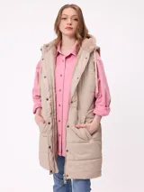 Chaleco largo acolchado color beige, con capucha forrada en sherpa, cierre frontal con cremallera y botones a presión, y bolsillos laterales.