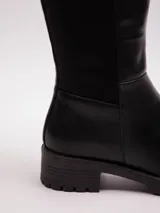 Bota de caña alta color negro, de símil cuero con pieza elástica en la parte trasera de la caña y cierre lateral. Cuenta con plataforma dentada.