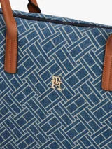 Cartera tote Tommy Hilfiger confeccionada en tejido denim azul con estampado geométrico de monogramas. Cuenta con asas superiores de cuero sintético marrón, cierre superior de cremallera y herrajes metálicos dorados con el monograma de la marca en el frente.
