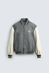 Cazadora estilo varsity o bomber, cuerpo principal gris oscuro y mangas color crema. Presenta cuello, puños y bajo en rib elástico con rayas grises y blancas. Tiene bordados en el pecho: un monograma estilizado a la izquierda y el texto "EST. 1975" a la derecha.