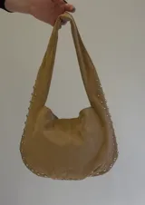 Cartera tipo hobo de gamuza color marrón claro, con detalles de tachas plateadas en los bordes y las asas. Cuenta con bolsillo interno con cierre.