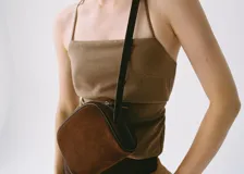 Bolso bandolera de tamaño pequeño confeccionado en piel de serraje color marrón. Cuenta con cierre de cremallera, forro interior de algodón y asa larga ajustable con hebilla metálica.