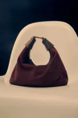 Cartera tipo hobo de Lolita, confeccionada en gamuza color bordó, con diseño de asa integrada y cierre superior. Presenta detalles en cuero liso en los extremos del asa y logo de la marca grabado en el frente.