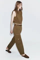 Pantalón cargo de tiro alto en color marrón tostado, con diseño de pernera holgada que se estrecha hacia el tobillo. Presenta bolsillos frontales con cierre de cremallera, bolsillos laterales de plastrón con solapa y botón, y cierre frontal con cremallera y botón.