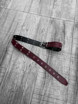 Cinto de cuero sintético color borgoña con hebilla metálica plateada y ojales metálicos plateados.