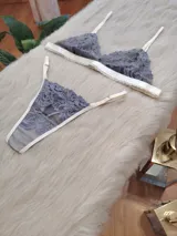 Conjunto de lencería compuesto por un corpiño triangular y una bombacha tipo tanga, confeccionados en encaje gris con detalles de terminaciones en color champagne.