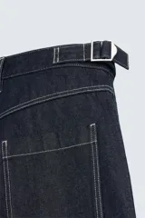 Bermuda de denim rígido color azul oscuro (sin prelavar) con corte amplio (baggy fit). Presenta pliegues frontales, trabillas ajustables en la cintura, bolsillos delanteros y bolsillos traseros de parche, todo resaltado con pespuntes a contraste.