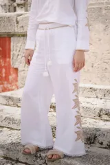 Pantalón blanco de lino, de pierna recta y corte holgado, con cintura elastizada, cordón ajustable con pompones y detalle de bordado lateral en tonos neutros. Presenta terminación desflecada en el bajo.
