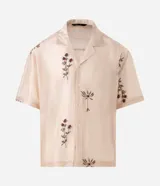 Camisa de lyocell color beige con corte boxy, cuello bowling y mangas cortas. Presenta un delicado bordado floral artesanal y cierre frontal con botones.