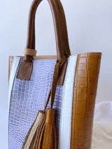 Bolso tipo tote de cuero color lila con textura croco, detalles en cuero marrón y laterales con franjas blancas. Incluye manijas de cuero marrón y accesorio de borlas colgantes.
