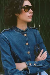 Chaqueta de denim azul con cuello redondo, manga larga y cierre frontal con botones dorados. Presenta costuras verticales decorativas y puños con botones a juego.