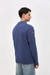 Buzo de punto color azul, con cuello alto y cierre frontal parcial. Presenta un diseño con textura acanalada en los hombros y mangas, y un pequeño bordado en la manga derecha.