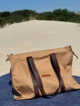 Bolso tote color beige con asas de cuero marrón, cierre superior y bolsillo interno.