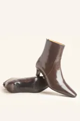 Bota de caña corta confeccionada en cuero vacuno color chocolate con acabado brillante. Presenta punta fina, cierre posterior y taco chupete bajo.