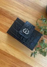 Tarjetero negro con textura símil cocodrilo, forro textil rojo y cierre con broche metálico plateado con diseño de venado.