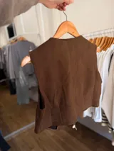 Chaleco de gamuza sintética color marrón, con escote en V y cierre frontal mediante lazos ajustables. Presenta detalles decorativos de tachas metálicas y apliques brillantes en el frente, además de dijes colgantes en los lazos.