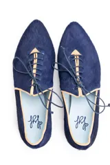 Zapato tipo blucher de pelo pony en color azul marino, con cierre de cordones y ribetes en color ocre. Presenta una silueta geométrica con aberturas laterales y tacón forrado en forma de lágrima.